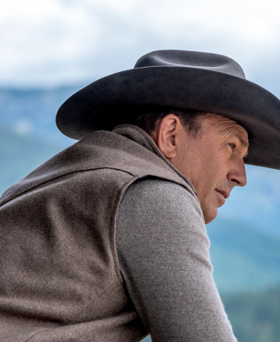 John Dutton wearing a cowboy hat and vest with a mountainous background