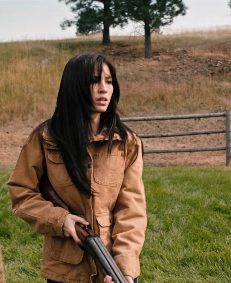 kelsey Asbille Yellowstone brown jacket holding a rifle in a grassy field with a wooden fence and trees in the background.
