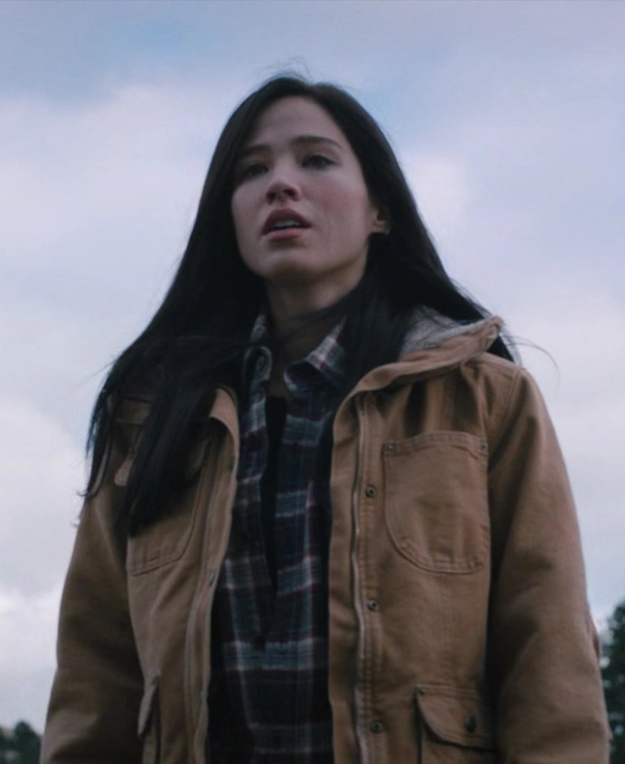 Monica wearing a brown jacket and plaid shirt outdoors with a cloudy sky.
