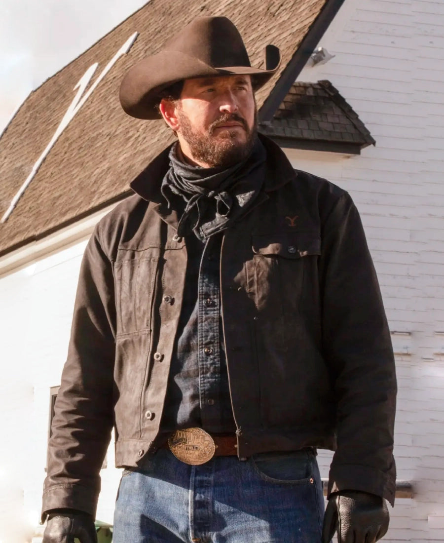 Rip Wheeler wearing a cowboy hat, leather jacket, and jeans standing in front of a Yellowstone building.
