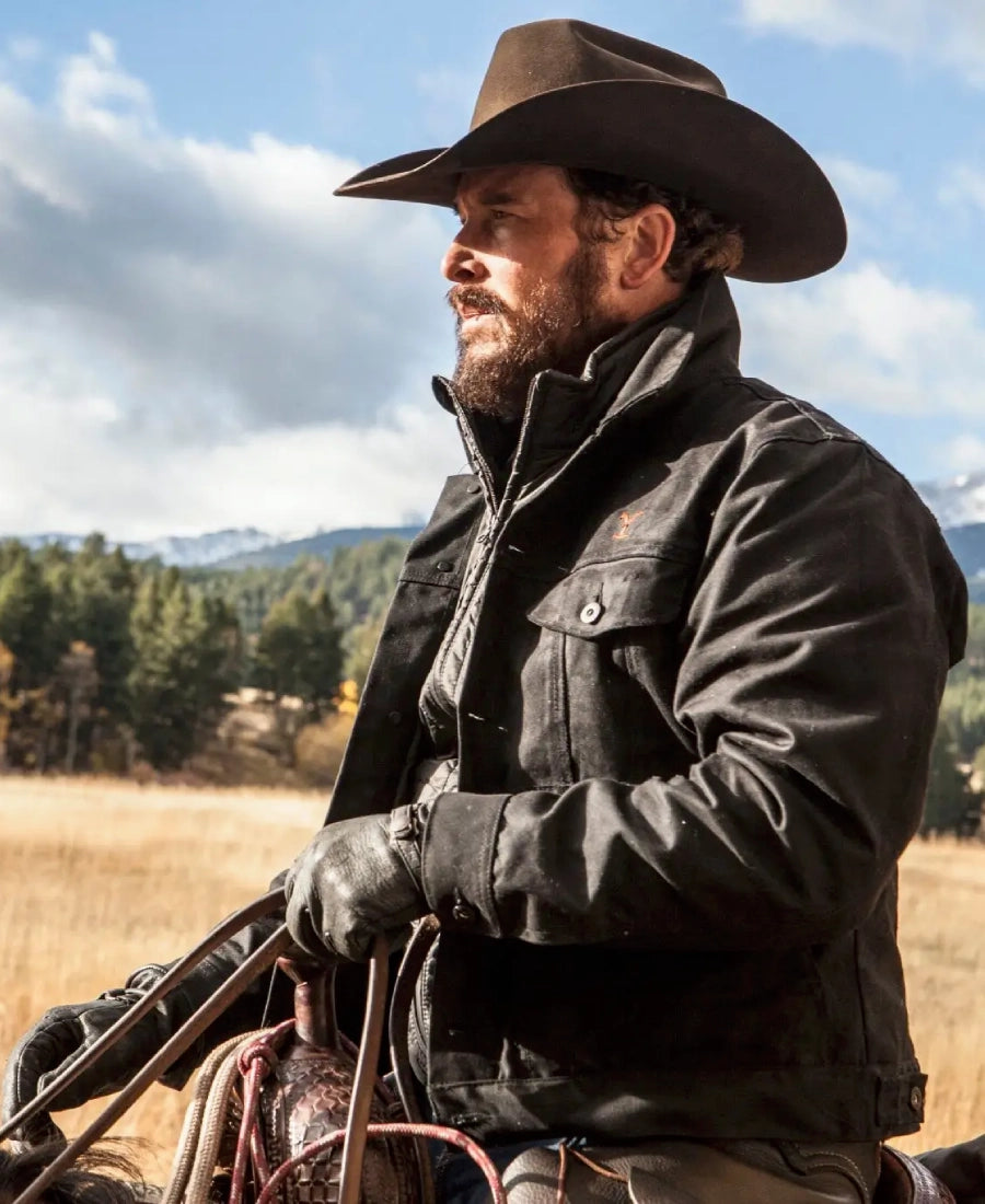in Yellowstone Rip Wheeler in a cowboy hat and leather jacket standing in a field with mountains in the background