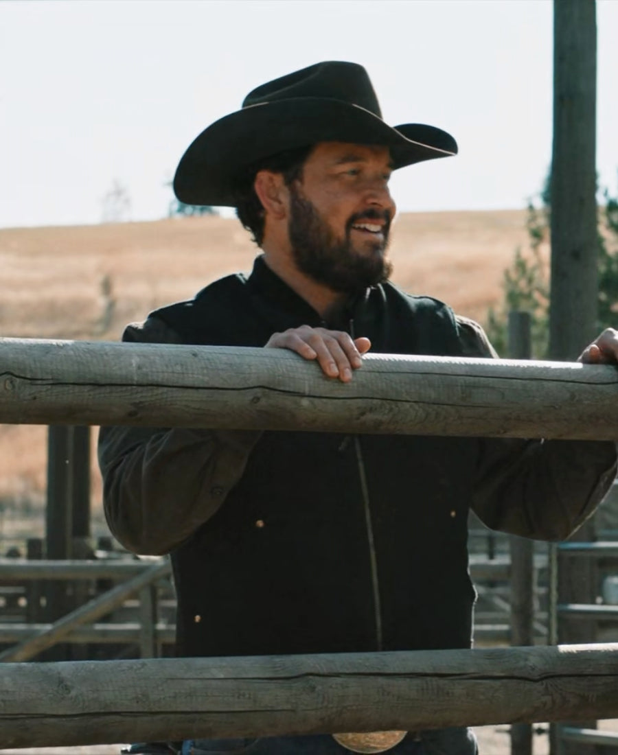Rip Wheeler Wool Vest wearing a black cowboy hat and jacket standing behind a wooden fence.