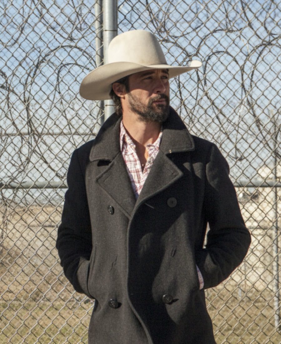 Walker Yellowstone Ryan Bingham wearing a dark coat and white cowboy hat standing in front of a chain-link fence.