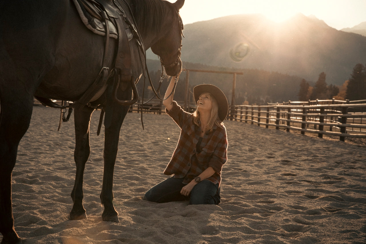 Yellowstone Beth Dutton in a cowboy hat kneeling next to a horse in a sandy area with mountains in the background