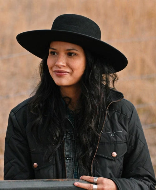 Yellowstone Avery Woman wearing a black hat and coat against a wooden background