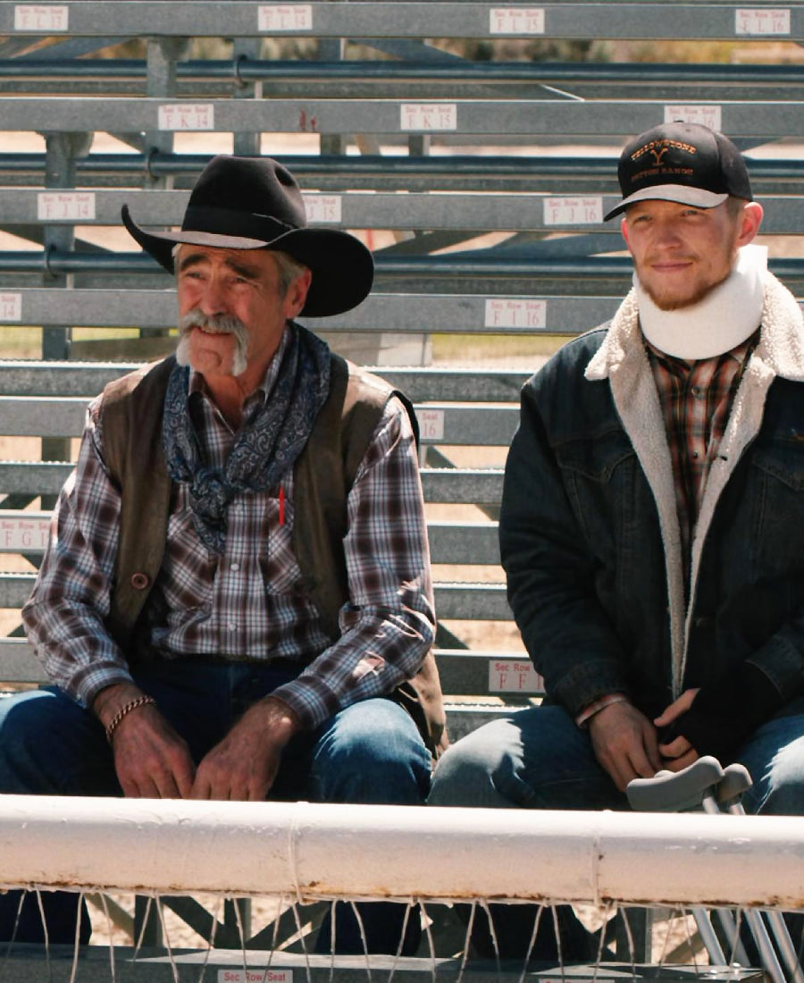Two men sitting on bleachers, one wearing a cowboy hat and plaid shirt, the other in a dark jacket with a white scarf.