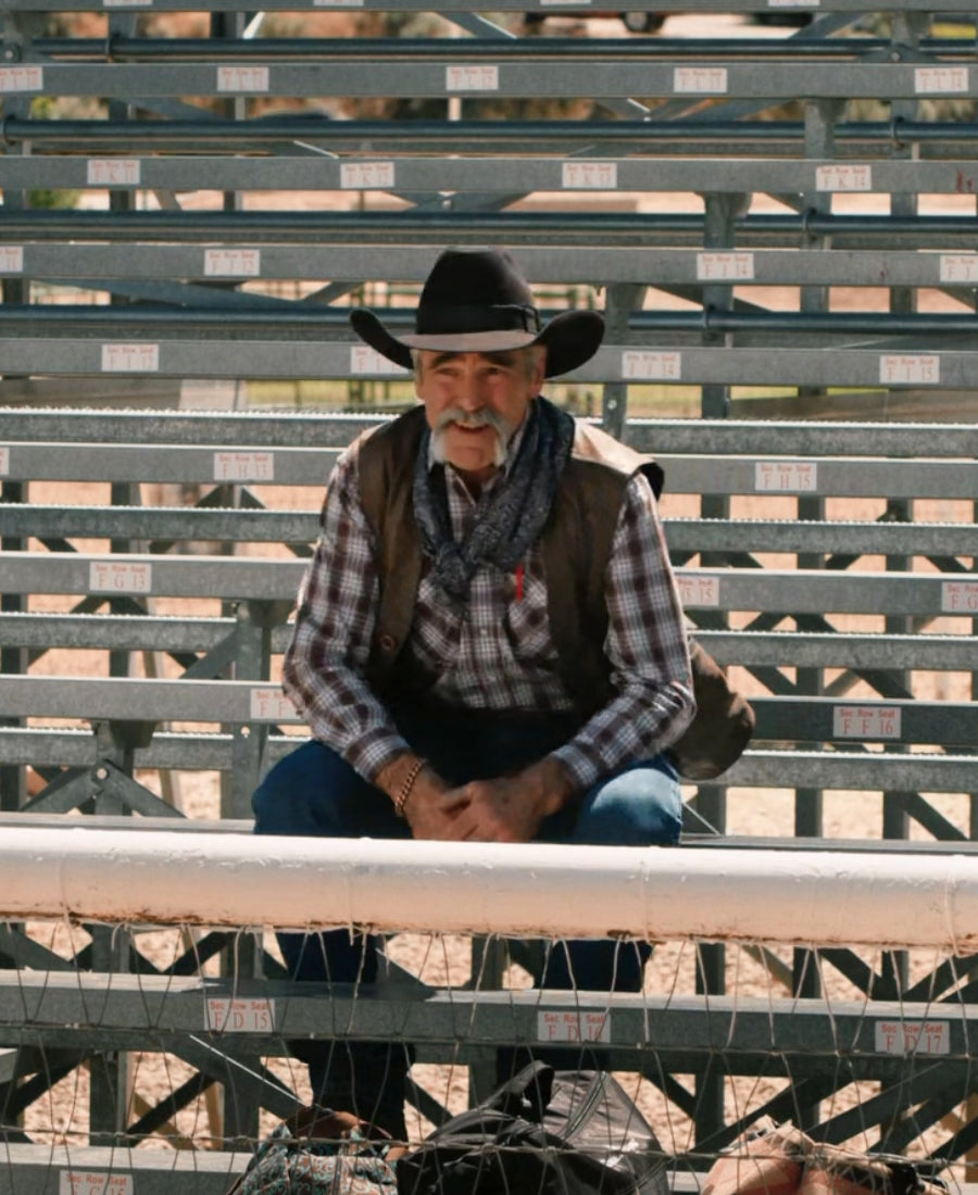 Yellowstone S04 Lloyd Vest Man in cowboy hat and plaid shirt sitting on metal cattle pens.