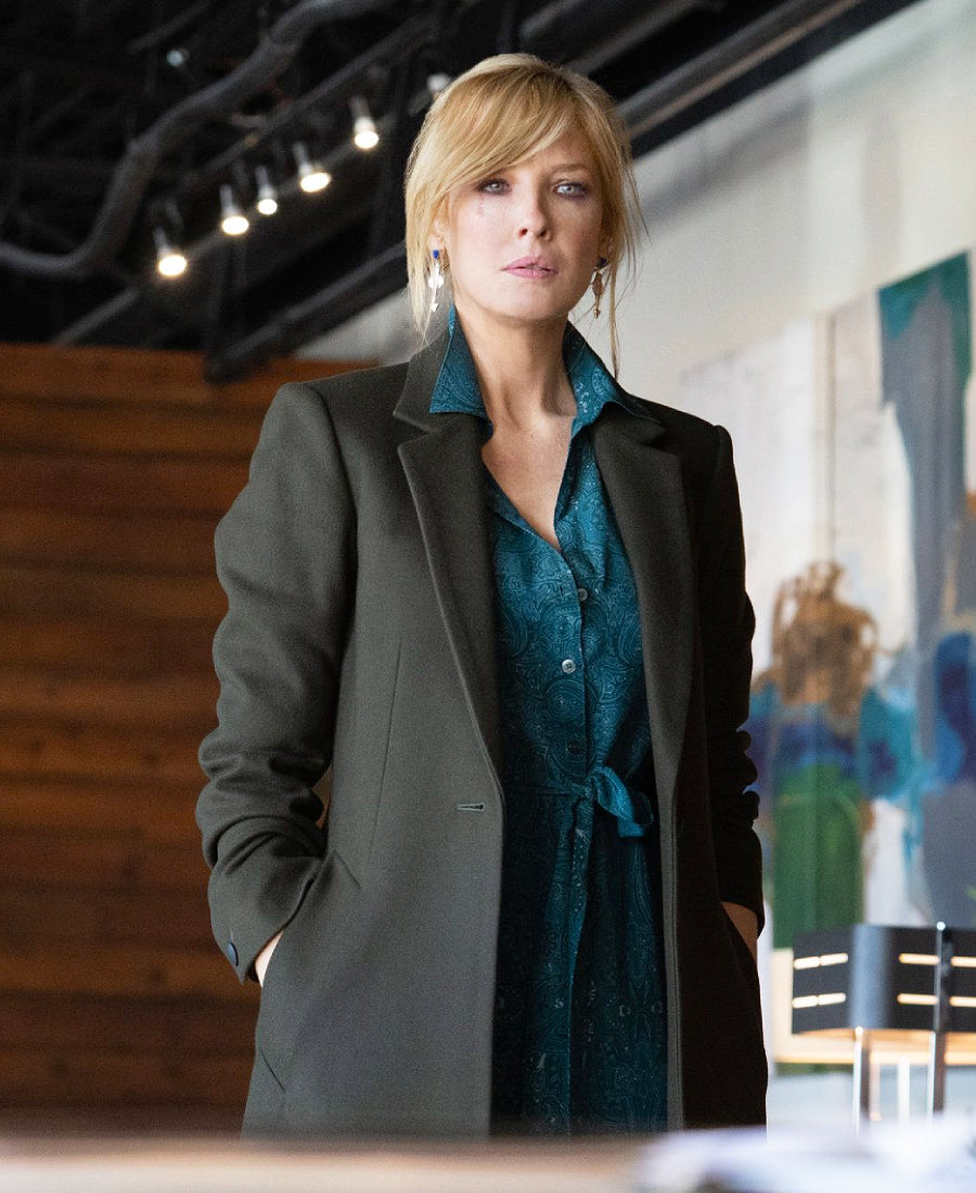 Beth Dutton wearing a gray coat over a teal shirt in an indoor setting