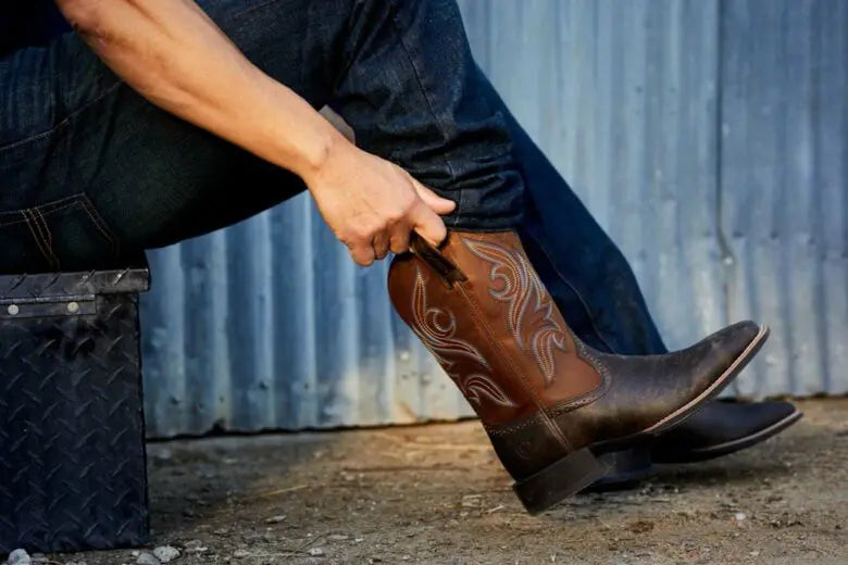 Person putting on a brown cowboy boot with white embroidery against a corrugated rip wheeler boot