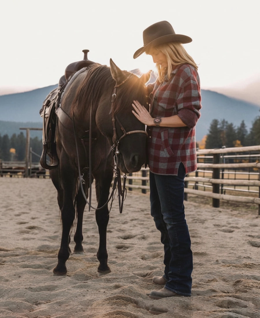 Beth Dutton wear a blazer coat and cowboy hat standing next to a horse in an outdoor setting with mountains in the background.