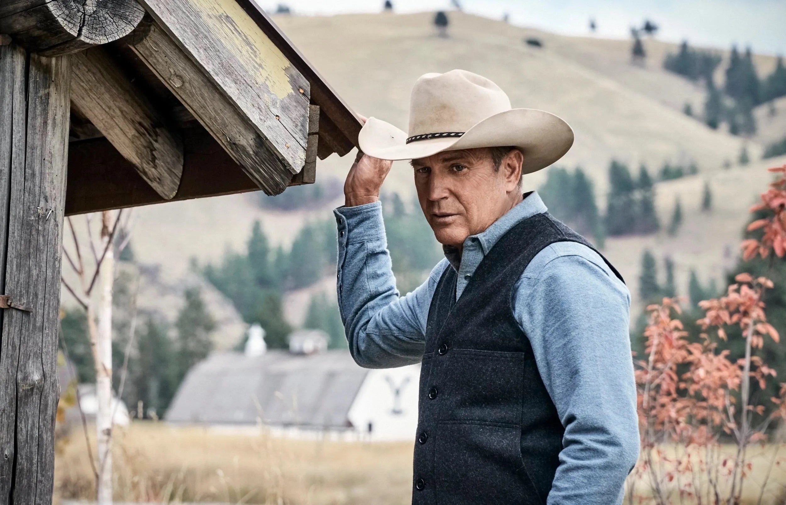 Man wearing a cowboy hat in a rural setting with mountains and trees in the background
