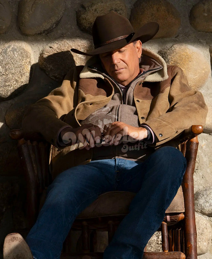 John Dutton wearing a brown jacket and cowboy hat sitting on a wooden chair against a stone wall.