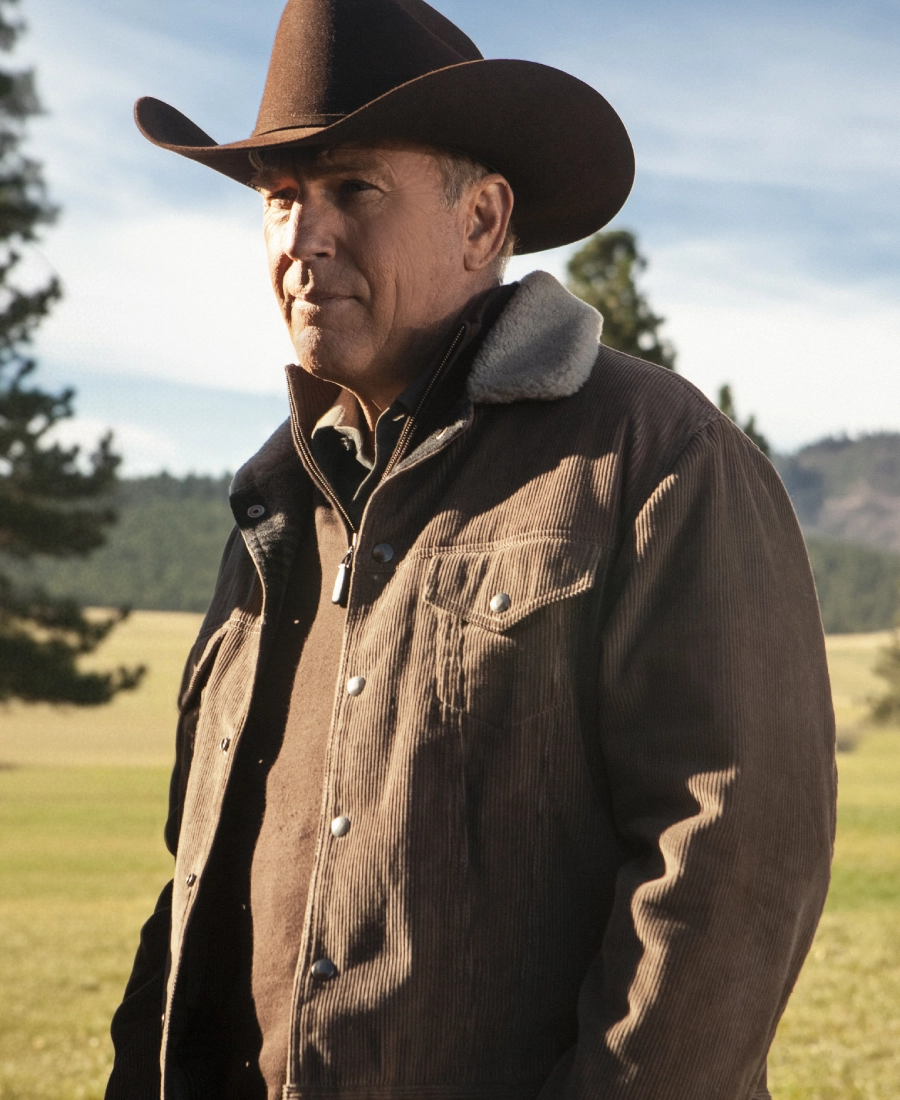 john Dutton Yellowstone wearing a brown cowboy hat and jacket in a natural setting with trees and mountains.