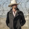 Walker Yellowstone Ryan Bingham wearing a dark coat and white cowboy hat standing in front of a chain-link fence.