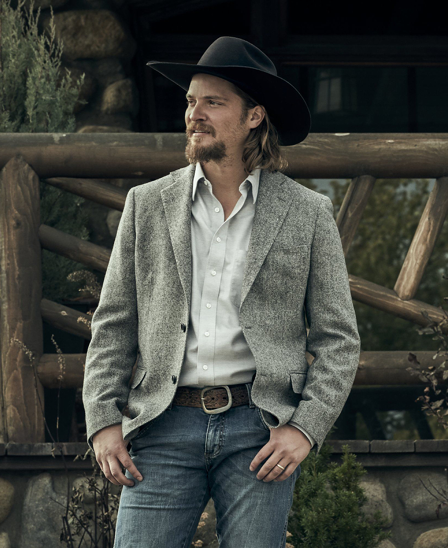 Kayce Dutton wearing a cowboy hat and gray blazer standing in front of a wooden fence.