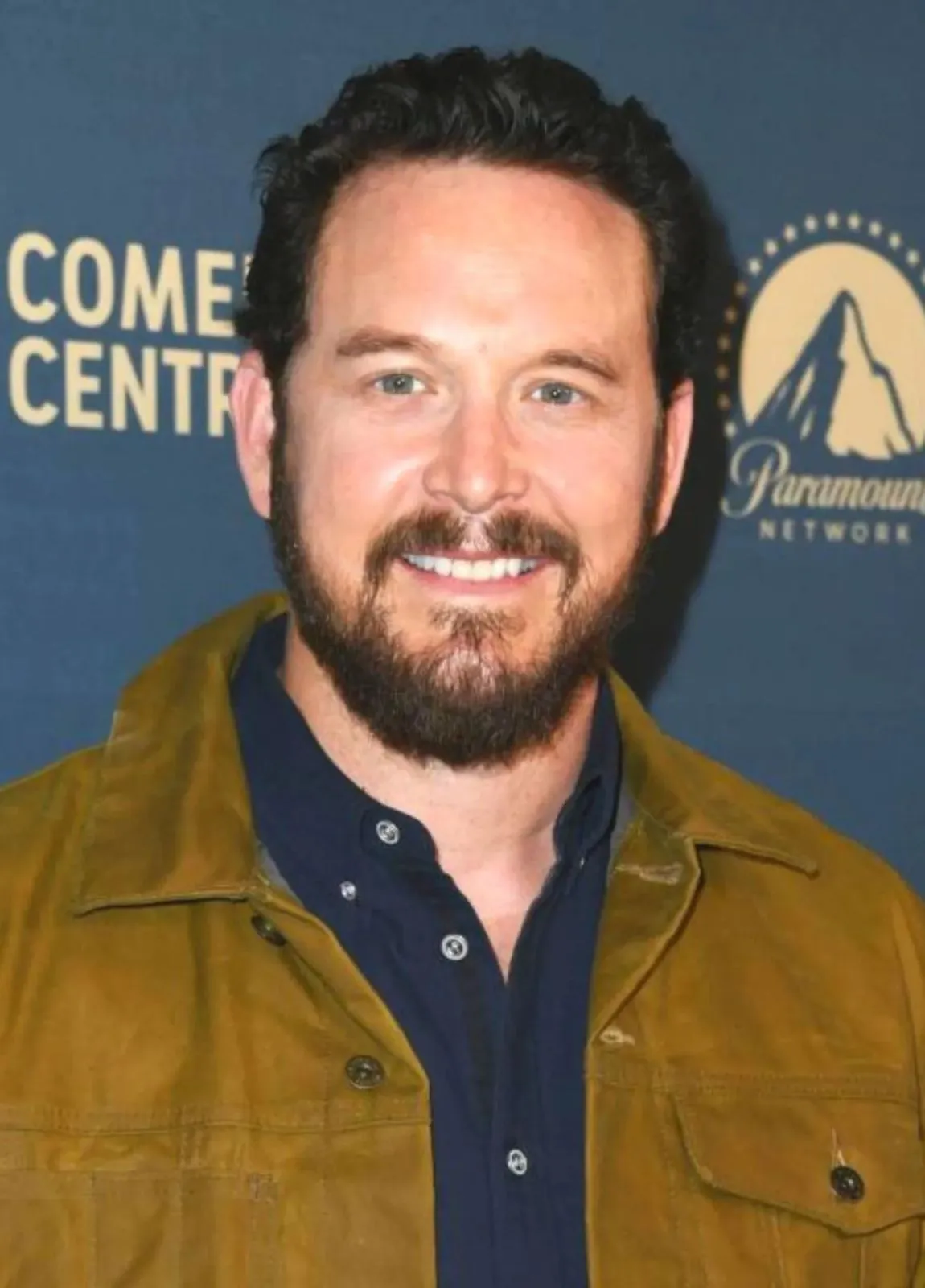 Cole-Hauser with a beard wearing a mustard jacket in front of a blue background with logos.