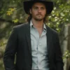 Kayce Dutton wearing a black hat and dark suit with a light shirt, standing outdoors with greenery in the background.