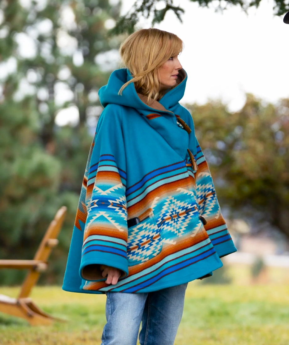 Beth Dutton wearing a blue patterned poncho outdoors with trees in the background