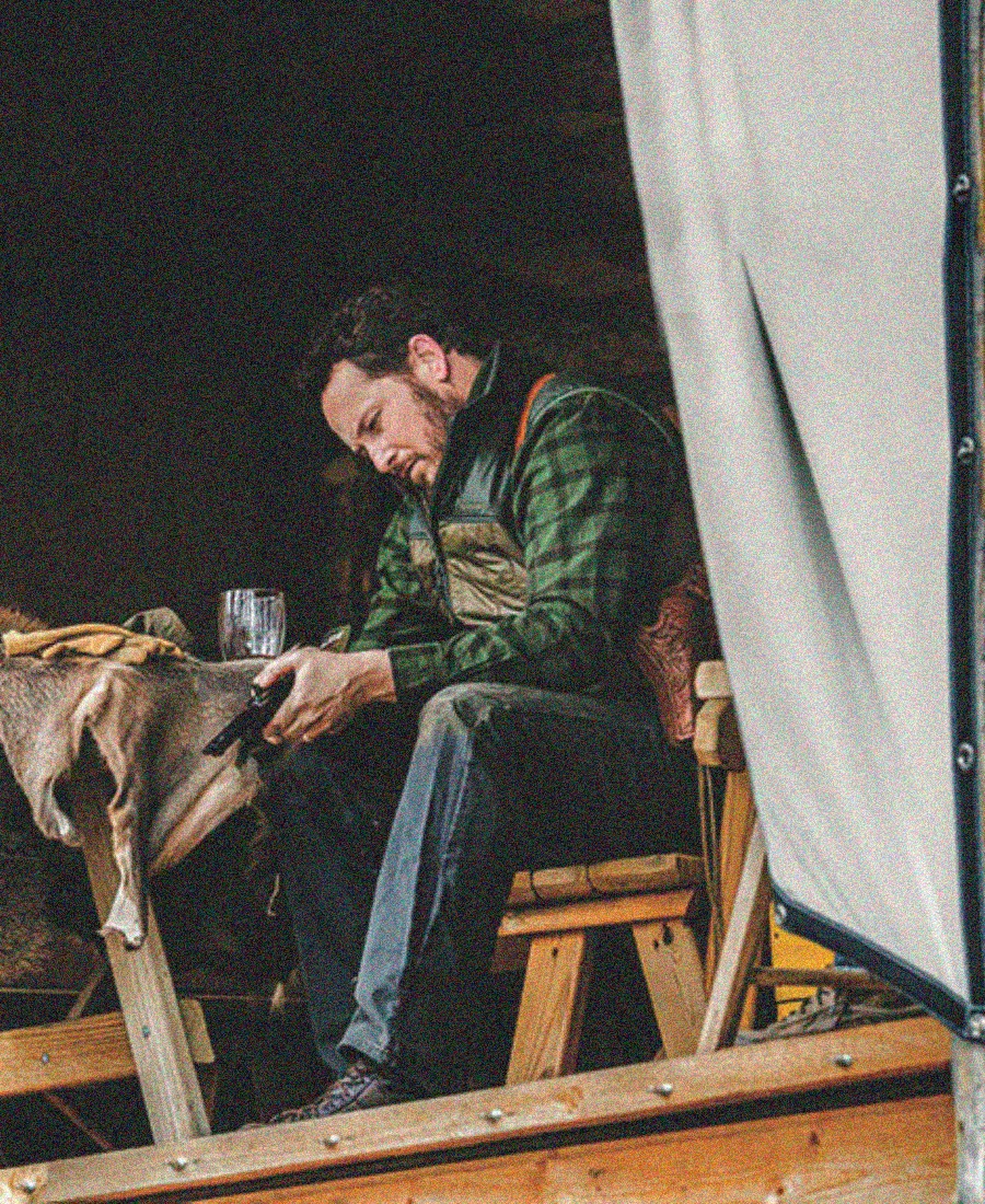 Rip Wheeler sitting at a wooden table in a rustic setting, wearing a green vest.