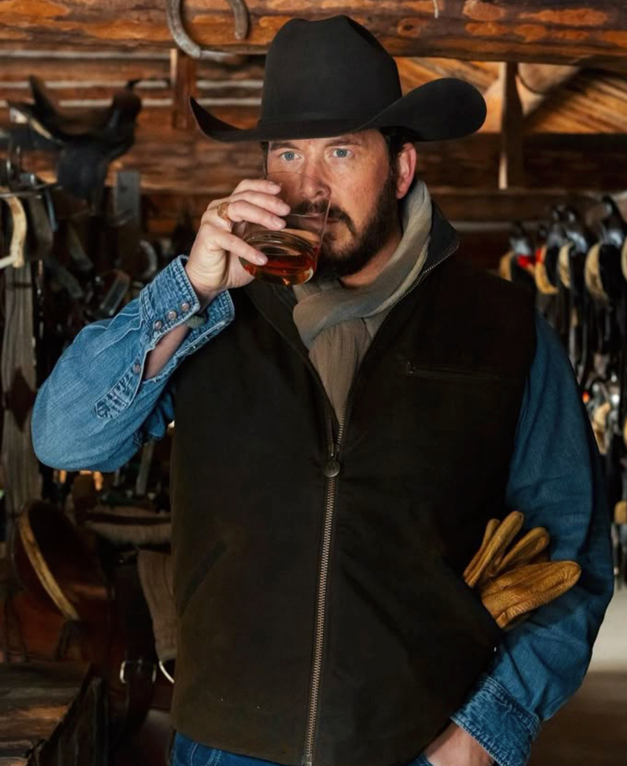 Rip Wheeler Black Wool vest with cowboy hat drinking from a glass in a rustic setting with wooden walls and horse equipment.