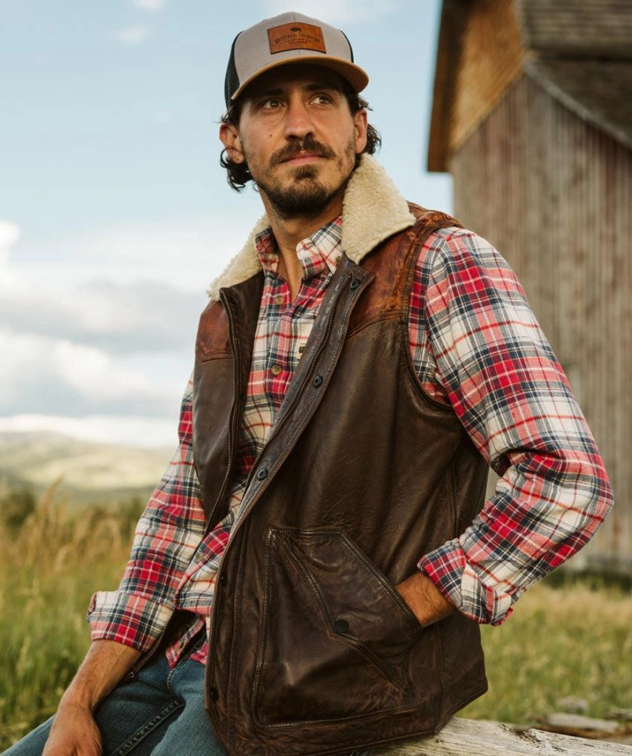 Man wearing a plaid shirt, brown vest, and cap outdoors with a wooden building in the background.