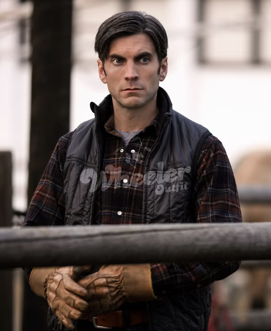 Jamie dutton wearing a plaid shirt and leather jacket with text, standing outdoors.