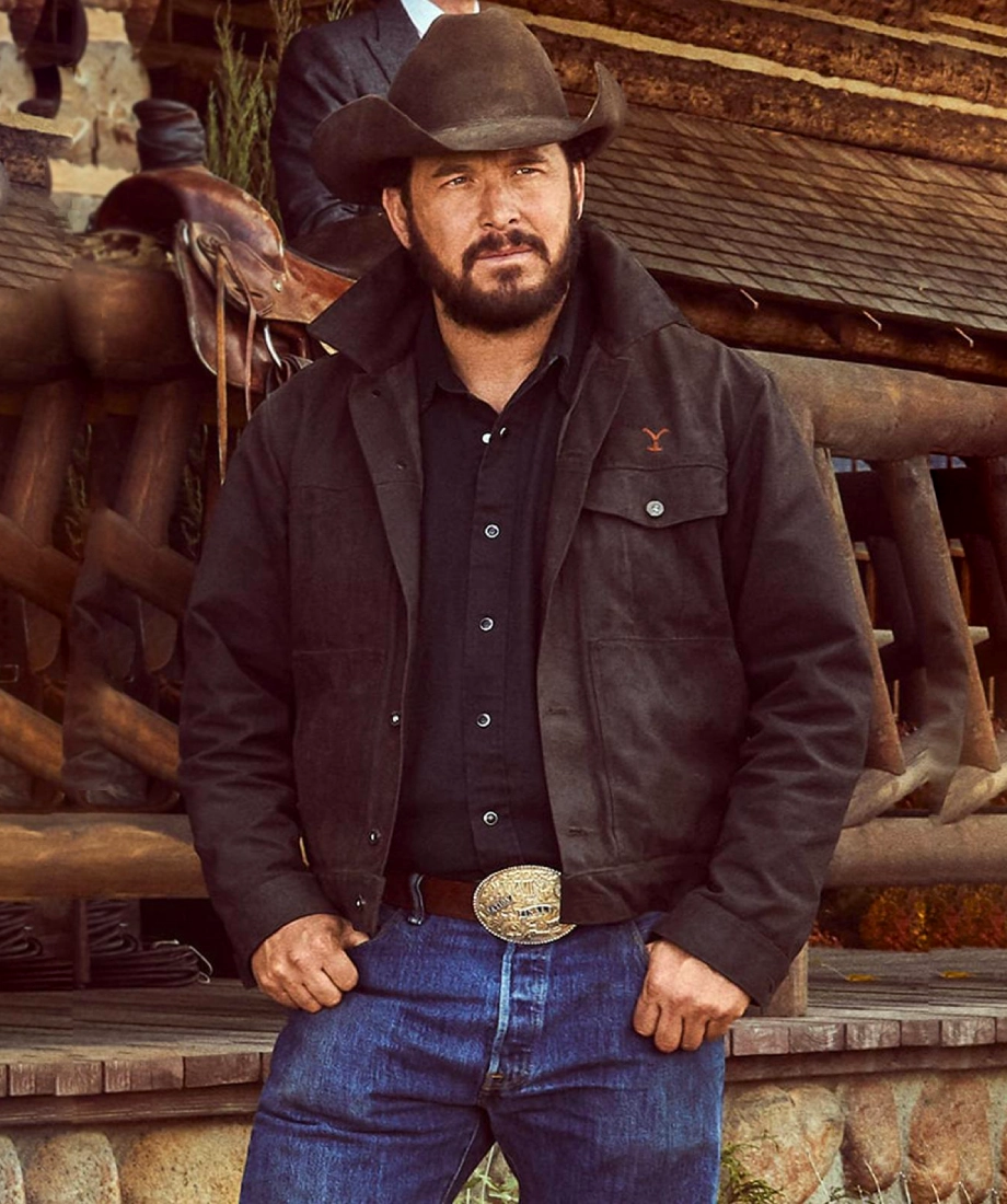 Rip Wheeler in cowboy hat and black cowboy jacket standing in front of wooden structures and a horse.
